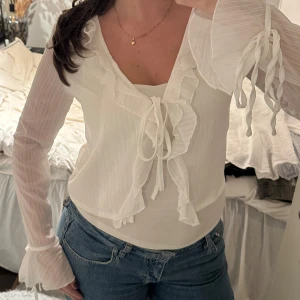 Vit blus med volanger och snörning - Säljer en söt vit blus med volangdetaljer och snörning framtill samt vid ärmsluten. Blusen har långärmade, lätt transparenta ärmar med diskret randigt mönster och v-ringad hals. Köpt i Spanien i storlek xs/s, oanvänd🤍