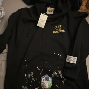 Svart hoodie Gallery Dept XL - Svart hoodie från Gallery Dept. i storlek XL med gul text 'DÉPT. de la GALERIE' på bröstet och konstnärliga färgstänk på fickan. Har snörning vid huvan och en patch på ärmen med texten 'Art on Display Courtesy of Josué Thomas'. Perfekt för dig som gillar streetwear.