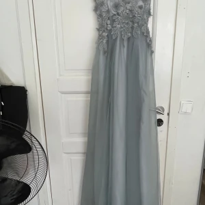 Ljusgrön långklänning med spetsdetaljer - Drömmig ljusgrön långklänning med tunna axelband och vacker blommig spets upptill. Kjolen är i flera lager tunn tyll som ger ett svävande intryck. Perfekt för bal eller student. Omlottkänsla och fina 3D-blommor på överdelen.