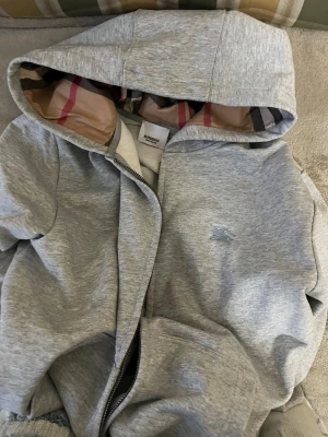 Grå hoodiejacka från Burberry - Säljer en grå hoodiejacka från Burberry med klassiskt rutigt foder i luvan och broderad logga på bröstet. helt Jackan har dragkedja med Burberry-detalj och är tillverkad i mjuk bomull och polyester. Perfekt för dig som gillar stilren design med lyxig touch.