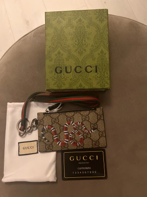 GG plånbok med orm och GG-mönster - Snygg plånbok från GG med klassiskt GG-mönster i beige och brunt, dekorerad med en cool röd, vit och svart orm på framsidan. Plånboken har dragkedja, flera kortfack och en grön-röd rem. Kommer med original ask och dustbag.