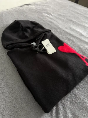 Ami Paris tröja  - Svart hoodie från Ami Paris Mattiussi med stor röd hjärtlogga och bokstaven A på bröstet. Tröjan har huva med snörning, ribbade muddar och känguruficka. Materialet är mjukt och stickat, perfekt för dig som gillar stilrena detaljer med en twist.