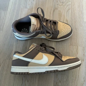 Nike Dunk Low i beige och brunt - Nike Dunk Low sneakers i beige och brunt med vit swoosh och bruna snören. Skorna har en klassisk låg siluett, perforerad tå och vit mellansula. Tillverkade i skinn med detaljer i syntet. Perfekta för dig som gillar stilrena och trendiga sneakers.