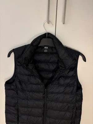 Svart dunväst från Uniqlo - Säljer en svart, quiltad dunväst från Uniqlo i storlek S. Västen är ultralätt och har dragkedja framtill samt hög krage. Perfekt för lager-på-lager och chill stil. Materialet är syntet och dun,