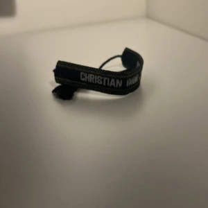 Christian Dior J'Adior svart armband - Passar alla storlekar. Snyggt svart armband från Christian Dior med texten 'J'Adior' broderad i vitt. Armbandet har guldfärgade detaljer och tofsar i ändarna för en cool touch. Perfekt accessoar för att lyfta din outfit med en lyxig känsla. 