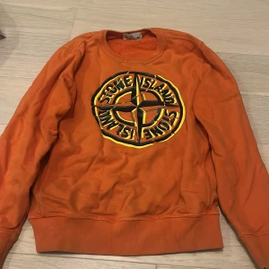 Orange sweatshirt från Stone Island Junior - Säljer en orange sweatshirt från Stone Island Junior med stort tryckt logomärke på bröstet i svart och gult. Tröjan har rund halsringning och ribbade muddar vid ärmslut och nederkant. Perfekt för dig som gillar streetwear och vill sticka ut.