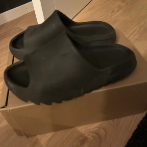 Svarta Yeezy Slide - Säljer ett par svarta Yeezy Slide sandaler från adidas. De har en chunky, räfflad sula och är tillverkade i mjukt syntetmaterial för maximal komfort. Perfekt för dig som gillar minimalistisk och trendig design. Box medföljer.