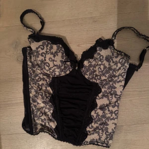 Svart och beige spetskorsett med mönster - Säljer en snygg korsett i svart och beige med blommigt spetsmönster och prickar. Toppen har reglerbara axelband, kupor med spetskant och dekorativt rynkparti framtill. Baksidan har hakknäppning och hela korsetten är detaljerad med spets och mesh.