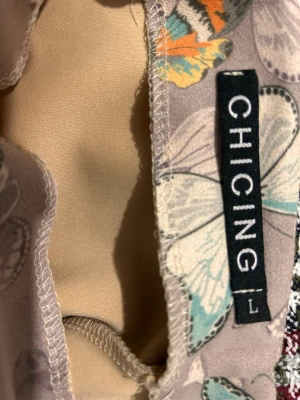 Kort kjol med fjärilsmönster från Chicing - Säljer en snygg kort kjol från Chicing med ett färgglatt fjärilsmönster i orange, turkos, svart och beige på en ljuslila bakgrund. Kjolen har en rak passform och är tillverkad i ett mjukt syntetmaterial som känns skönt mot huden. Perfekt för dig som gillar unika prints!