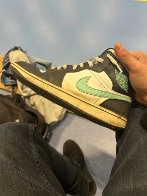Nike Air Jordan 1 svart/vit mint - Nike Air Jordan 1 sneakers i svart och vit med mintgrön swoosh och Air Jordan-logga på sidan. Klassisk high-top siluett med snörning och perforerad tå. Skorna är tillverkade i skinn och har en platt sula. Perfekt för dig som gillar streetwear och vill sticka ut.