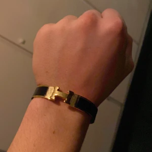  svart och guld armband - Snyggt armband i svart och guld med den ikoniska H-detaljen framtill. Armbandet är i metall med en blank finish och har en stilren, modern design som verkligen sticker ut. Perfekt accessoar för dig som gillar lyxiga och klassiska smycken.