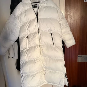 Vit pufferjacka från Schott NYC XL - Schysst vit pufferjacka från Schott NYC i storlek XL. Jackan har huva, två dragkedjeförsedda fickor fram och en snygg logga på ärmen. Den är vadderad och har en clean look med breda paneler och dragkedja framtill.