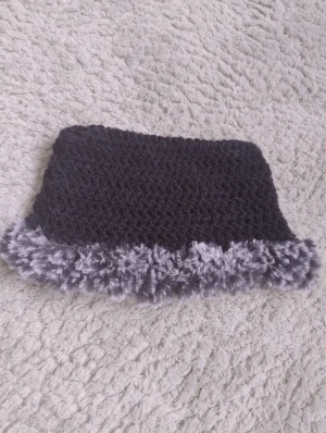 Svart handgjord virkad mössa med pälsdetalj - Säljer en svart virkad mössa med fluffig grå pälsdetalj runt kanten som jag gjort själv. Mössan har en fyrkantig form som får det att se ut som att man han kattöron när man har på sig den. Perfekt för dig som vill sticka ut och hålla dig varm under kalla dagar.
