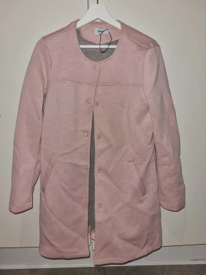 ONLY lång cardigan jacka i tre färger - Snygg och stilren lång cardigan jacka från ONLY med rund hals, tryckknappar och två fickor fram. Finns i rosa, beige och svart. Jackan har en clean look och är tillverkad i mjukt material som ger en chill vibe. Perfekt för dig som gillar minimalistisk stil.