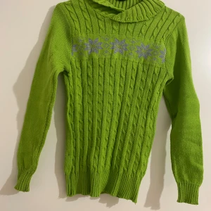 Grön polotröja med lila mönster - Snygg grön polotröja med kabelstickat mönster och lila snöflingor över bröstet. Tröjan har lång ärm, ribbade muddar och en bred, ribbad polokrage som ger extra mysfaktor. Perfekt för dig som gillar färg och vill sticka ut lite extra.