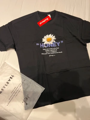 Svart t-shirt med blomtryck från Eastlevel - Svart t-shirt från Eastlevel med tryck av en stor prästkrage och texten 'HONEY' på bröstet. T-shirten har rund hals, korta ärmar och är gjord i mjuk bomull. Perfekt för dig som gillar streetstyle och coola prints.
