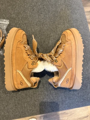 UGG sneakers med fuskpäls och platåsula - Bruna sneakers från UGG med chunky platåsula, breda mönstrade snören och vit fuskpäls på insidan. Ovandel i mocka och textil, detaljerad med UGG-logga på sidan. Perfekta för höst och vinter när du vill ha både stil och värme.