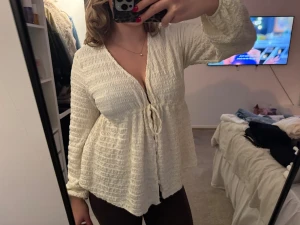 Vit omlottblus med knyt och lång ärm - Säljer en superfin vit omlottblus med knyt framtill och v-ringad hals. Blusen har ett smockat, strukturerat tyg som ger en cool och luftig känsla. Långa ärmar med lätt puff och flowig passform. Perfekt för dig som gillar romantisk och chill stil.