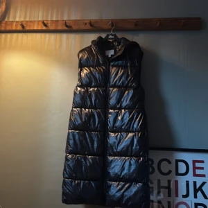 Svart lång dunväst från H&M - Säljer en svart, lång dunväst från H&M med huva och glansig yta, fickor på var sida. Västen har quiltad design och dragkedja framtill. Perfekt för lager-på-lager och extra värme under kyliga dagar. Snygg och enkel att matcha med hoodies eller stickade tröjor.