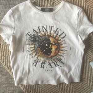 Vit croppad t-shirt med draktryck från Bershka - Säljer en vit croppad t-shirt från Bershka med coolt draktryck och texten 'Tainted Heart' på framsidan. Rund hals och korta ärmar, tillverkad i mjuk bomull. Perfekt för dig som gillar edgy och unik stil.