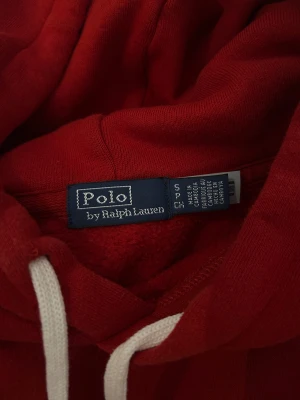 Röd hoodie från Polo Ralph Lauren - Snygg röd hoodie från Polo Ralph Lauren med vit snörning i huvan och klassisk blå logga broderad på bröstet. Tröjan har en stor känguruficka framtill och är tillverkad i mjuk bomull. 