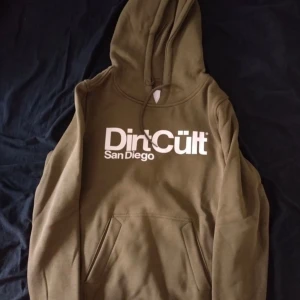 Olivgrön hoodie från DirtCult - Säljer en olivgrön hoodie från DirtCult San Diego med stor vit logga på bröstet. Tröjan har huva med snörning och en klassisk magficka. Perfekt för dig som gillar streetwear och vill ha en chill look.
