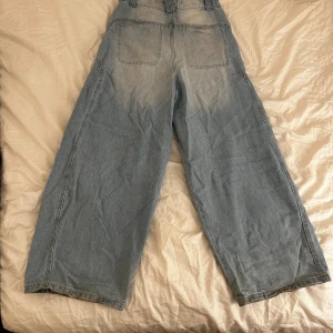 Wide jeans i ljusblå denim från Bershka - Säljer ett par ljusblå jeans från Bershka i storlek 36. Modellen har hög midja, raka och breda ben samt stora fickor bak. Jeansen är tillverkade i klassisk denim och har en avslappnad, trendig siluett som är perfekt för en chill streetstyle.