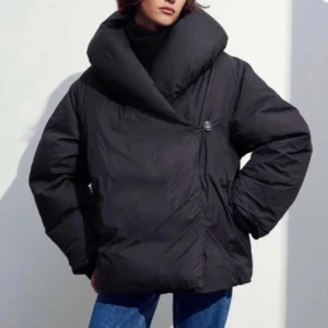 Svart pufferjacka med stor krage - Säljer en svart pufferjacka med oversized passform och stor, mysig krage. Jackan har en unik omlott-design med knappstängning på sidan och långa ärmar. Perfekt för dig som vill ha en cool och varm jacka med modern vibe.