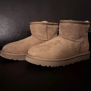 Beige mockaboots med ullfoder som likar uggs - Mysiga uggsliknande beige mocka med rund tå och platt sula. Insidan är fodrad med mjuk, ljus ull som håller fötterna varma. Snygga detaljer med grova sömmar och dragflik baktill. Perfekta för kalla dagar och chill stil. Liknar uggs rakt av förutom mönstret på sulan dock identisk när det kommer till sömmar färg och form och helt utan några loggor