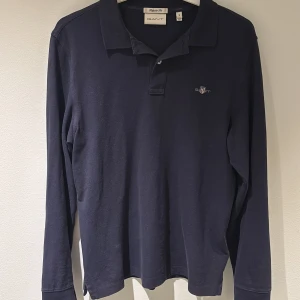 Marinblå långärmad pikétröja från GANT - Snygg marinblå långärmad pikétröja från GANT i regular fit. Klassisk krage med två knappar och broderad logga på bröstet. Tröjan är rätt i storleken och därmed passande för dig med S. Hör av dig vid funderingar.