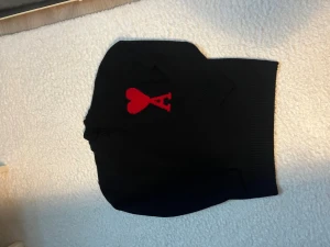 Svart stickad tröja med rött hjärta - Svart stickad tröja från AMI Paris med ett stort rött hjärta och A på bröstet. Tröjan har ribbade muddar vid hals, ärmslut och nederkant. Klassisk passform och mjukt material som känns skönt mot huden. Perfekt för dig som gillar stilrena detaljer. Storlek M 