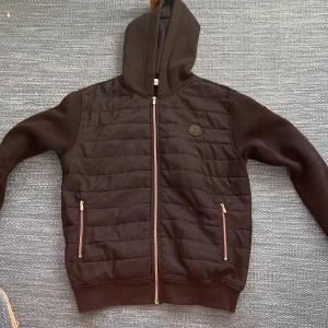 Svart quiltad pufferjacka Moncler - Svart pufferjacka från Moncler med huva, quiltad framsida och två fickor med dragkedja. Jackan har Moncler-logga på bröstet och ärmen, samt hel dragkedja framtill. Materialet är en mix av polyester och bomull, med ribbade muddar och långärmad passform. (Pris kan diskuteras)