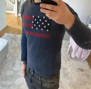 Lexington mörkblå stickad tröja  - Snygg mörkblå stickad tröja från Lexington med amerikansk flagga i rött och vitt på bröstet. Tröjan har rund hals, ribbade muddar och lång ärm. Perfekt för dig som gillar klassisk stil med en twist.