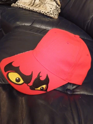 Ny Keps TENGTOOLS - Cool röd keps TENGTOOL med broderade gula och svarta ögon på skärmen. Kepsen har klassisk form med böjd skärm och ventilationshål upptill. Perfekt för dig som vill sticka ut med en unik och färgstark accessoar.