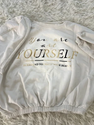 Vit sweatshirt med guldtryck Sinsay XS - Vit sweatshirt från Sinsay i storlek XS med snyggt tryck i guld och svart på framsidan. Tröjan har rund hals, långa ärmar med puff och resår vid ärmslut och nederkant för en cool och avslappnad look. Perfekt för dig som gillar statement-plagg.