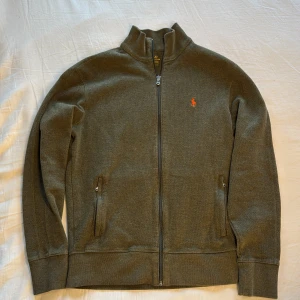 Grön ziptröja från Polo Ralph Lauren - Grön långärmad ziptröja från Polo Ralph Lauren med klassisk ståkrage och orange broderad logga på bröstet. Tröjan har två fickor med dragkedja och är tillverkad i mjuk bomullsmix som känns skön mot huden. Perfekt för chill dagar och enkel att matcha. Priset går att diskuteras vid snabbaffär!