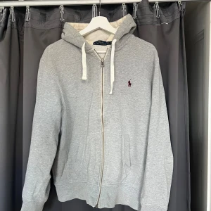 Grå hoodie från Polo Ralph Lauren - Snygg grå hoodie från Polo Ralph Lauren med dragkedja och vit snörning i huvan. Tröjan har ett broderat vinrött Polo-logo på bröstet, två fickor fram och mjukt bomullsmaterial. Perfekt för chill dagar och streetwear-vibe.