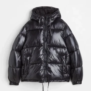 Svart pufferjacka med huva - Svart, glansig pufferjacka med huva och snörning. Jackan har dragkedja framtill, två sidofickor och elastiska muddar vid ärmslut. Den är vadderad och har en oversized look, perfekt för kalla dagar. Snygg och enkel att matcha med streetwear.