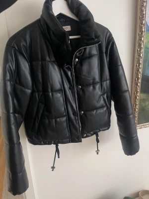 Svart croppad jacka  - Säljer en svart croppad pufferjacka från Nelly. Jackan har dragkedja och knappar framtill samt snörning i nederkant för att justera passformen. Jätte fint skick 