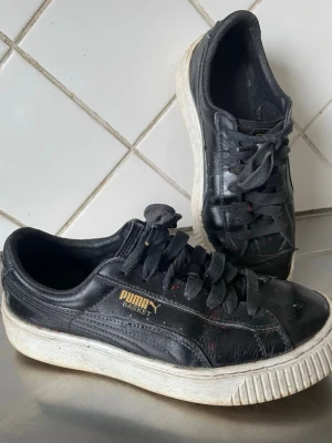 Svarta Puma Basket sneakers - Svarta Puma Basket sneakers med klassisk låg profil och vit platt sula. Skorna har snörning och är tillverkade i skinn med guldfärgad Puma-logga på sidan. Perfekt för dig som gillar retrostil och vill ha ett par tidlösa sneakers med cool vibe.