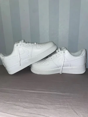 Nike Air Force 1  - Klassiska helvita Nike Air Force 1 sneakers i skinn med låg profil och rund tå. Snygg och clean design med snörning och Nike-logga på sidan och hälen. Perfekta till jeans eller shorts för en stilren look.