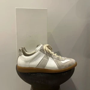 Maison margiela  - Snygga sneakers i beige och vitt med coola paneler i mocka och skinn. Dom har 2 små defekter som ni ser på bild 4  då det har lossnat lite vit färg men inget man ser eller tänker på vid andväning. Perfekt för dig som gillar stilrena och neutrala färger med en retrovibe. Äktihetsbevis finns! Dustbag tillkommer!