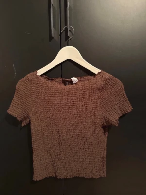 Brun ribbad croppad topp - Säljer en brun croppad topp från SHEIN med korta ärmar och ribbad struktur. Toppen har en lätt vågig kant vid halsen och är tillverkad i ett stretchigt syntetmaterial som sitter snyggt på kroppen. Perfekt att styla med jeans eller kjol.