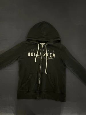 Svart zip hoodie från Hollister - Säljer en svart zip hoodie från hollister. Den har vit och grå text framtill och vita snören. Storlek S men skulle nog kunna funka som XS. Har varit en favorit för mig och har inga defekter förutom att den är lite nopprig, säljer eftersom den är för liten🥹💗 Priset är prutbart!!