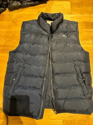 Mörkblå dunväst från GANT - Snygg mörkblå dunväst från GANT med hög krage och diskret logga på bröstet. Västen har quiltad design, två sidofickor med dragkedja och en kraftig dragkedja framtill. Perfekt att slänga över en hoodie för extra värme. Nypris 1.600kr.