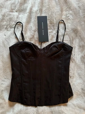 Svart korsettopp med leopardmönster från Bershka - Snygg svart korsettopp från Bershka i storlek S med tunna justerbara axelband och subtilt leopardmönster. Toppen har markerade kupor och sömmar som ger en cool siluett. Materialet är glansigt och känns lyxigt mot huden. Lappen är kvar