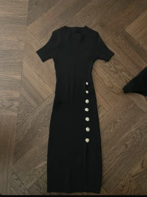 Svart ribbad långklänning med knappar - Snygg svart långklänning med ribbad struktur och korta ärmar. Klänningen har dekorativa silverfärgade knappar längs sidan och en liten slits nedtill. Perfekt för dig som gillar stilrena och trendiga plagg med coola detaljer.