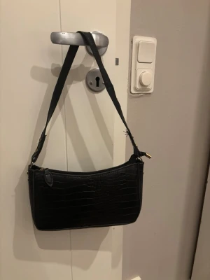 Svart axelväska i croco-mönstrat skinn - Snygg svart axelväska med croco-mönstrad yta och justerbar rem. Väskan har en stilren, rektangulär form och dragkedja upptill med gulddetalj. Perfekt storlek för det viktigaste och ger en cool vibe till din outfit.