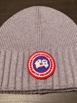Grå ribbstickad mössa från Canada Goose - Snygg grå mössa från Canada Goose med ribbstickad design och klassisk rund logotyp-patch framtill. Mössan är tillverkad i mjukt material och har en bred uppvikt kant för extra stilpoäng. Perfekt för kalla dagar när du vill hålla dig varm och se cool ut.