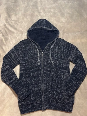 Stickad Cardigan - Säljer en grå stickad hoodie med mysigt fleecefoder i huvan. Tröjan har dragkedja framtill, snörning vid huvan och ribbade muddar. Perfekt för kyliga dagar och har ett snyggt melerat mönster som ger en cool vibe.